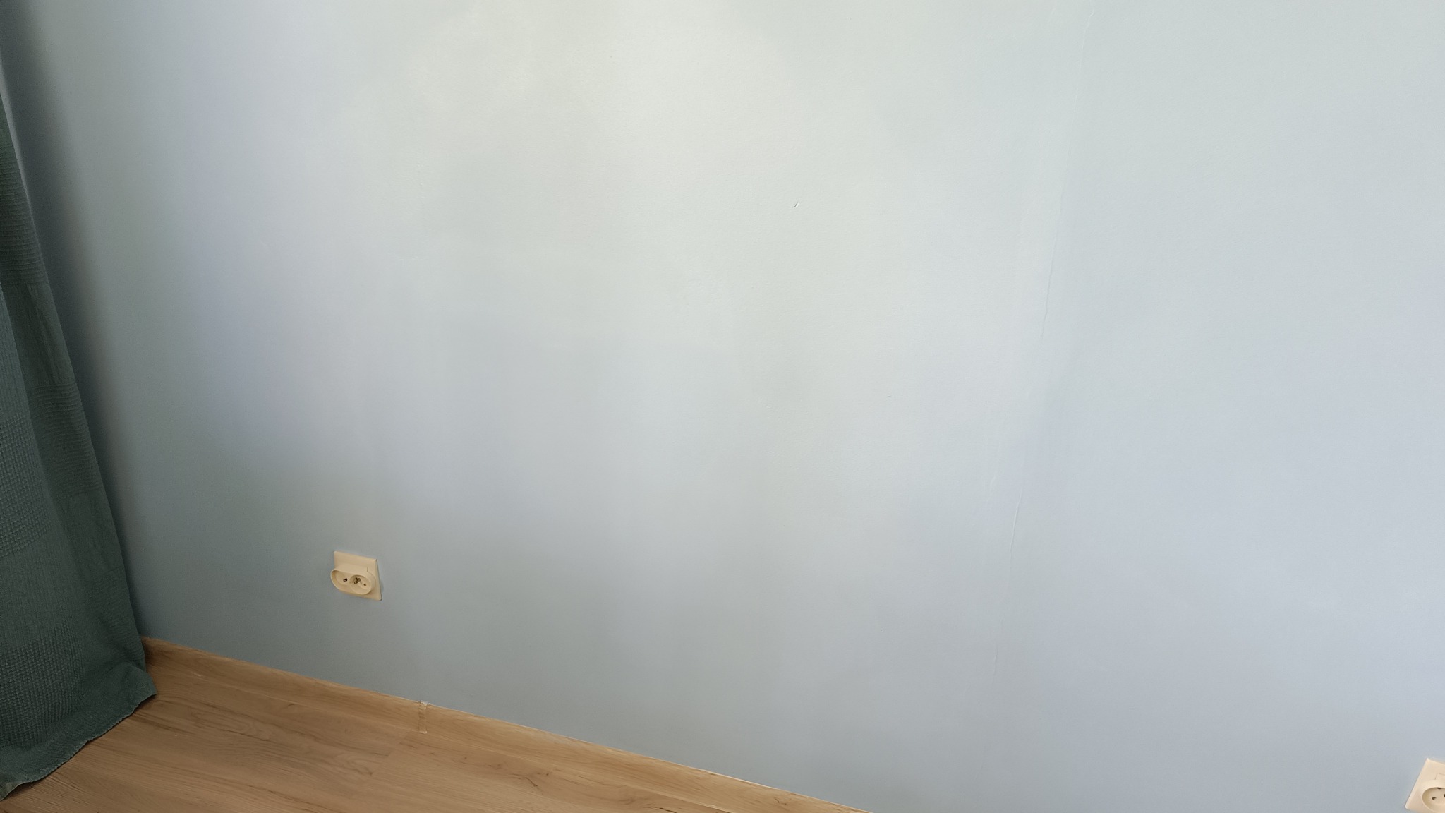 Ściana pomalowana na jasny odcień niebieskiego, widoczne dwa gniazdka elektryczne, fragment drewnianej podłogi i zasłony w lewym rogu.