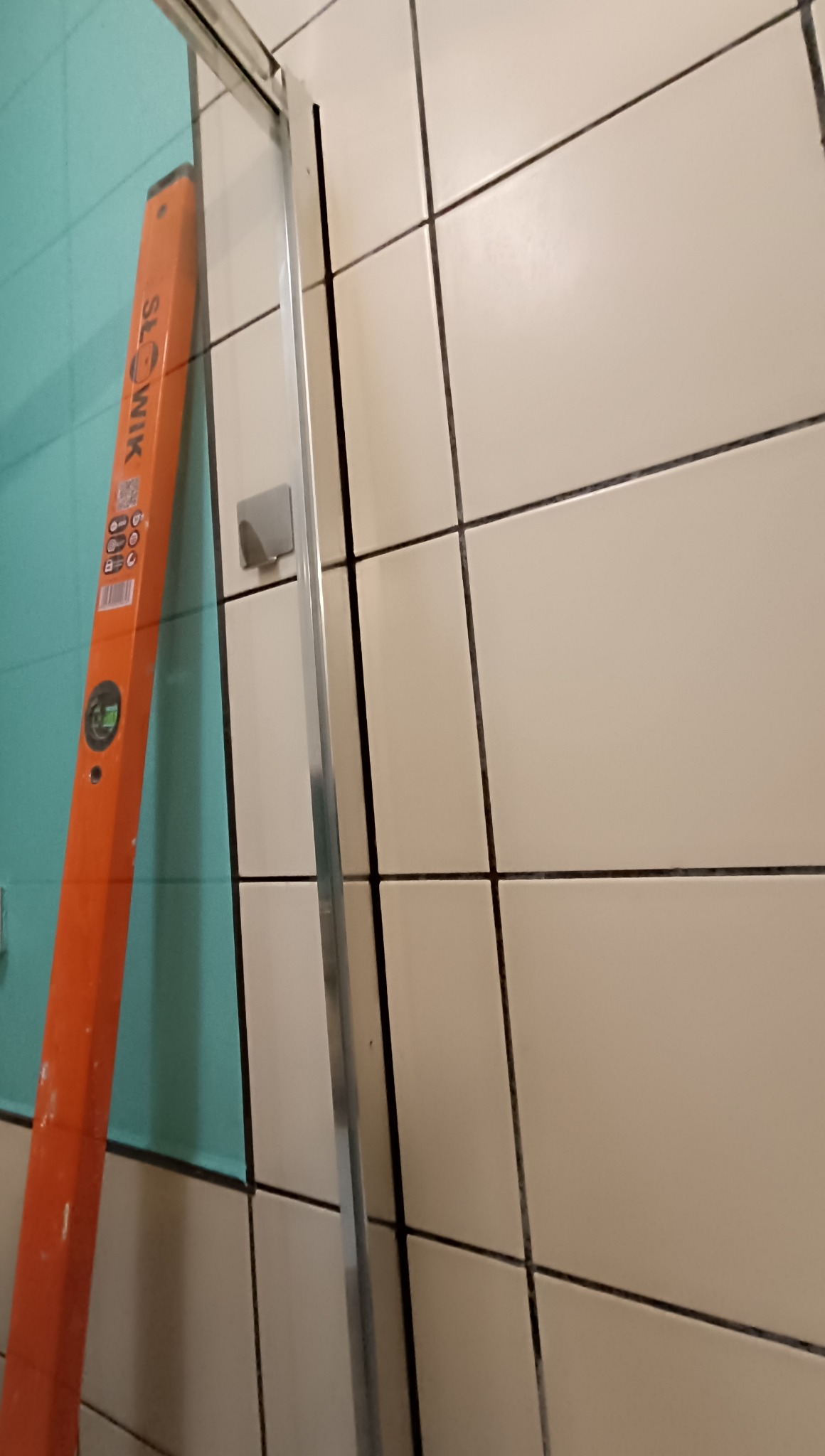 Białe i turkusowe płytki w łazience z poziomnicą i profilem aluminiowym, sugerujące montaż kabiny prysznicowej lub prace wykończeniowe.
