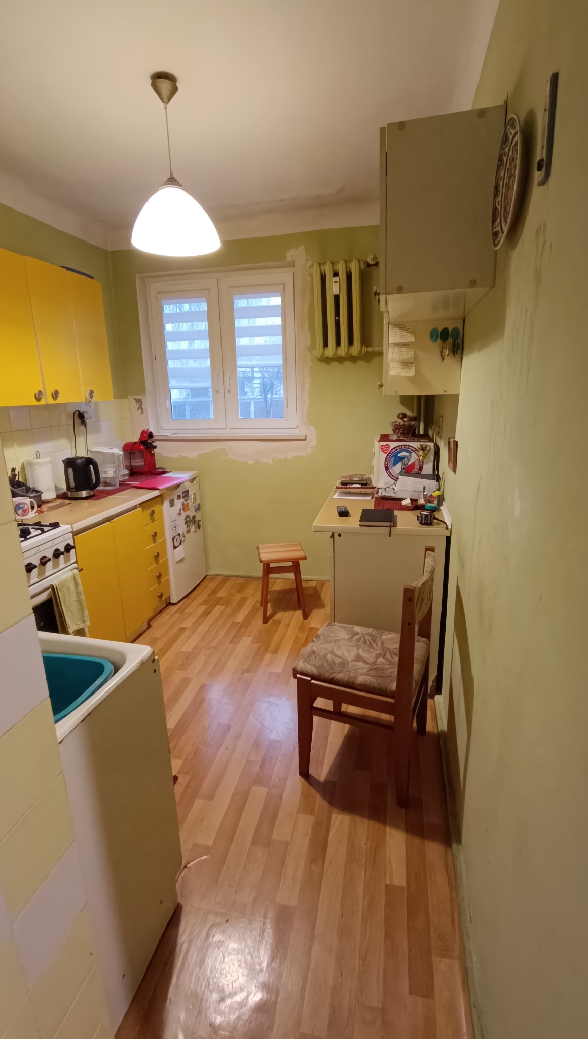 Kuchnia w trakcie odświeżania: żółte szafki, drewniana podłoga i ściany przygotowane do malowania wokół okna. Widoczne meble i sprzęty kuchenne.