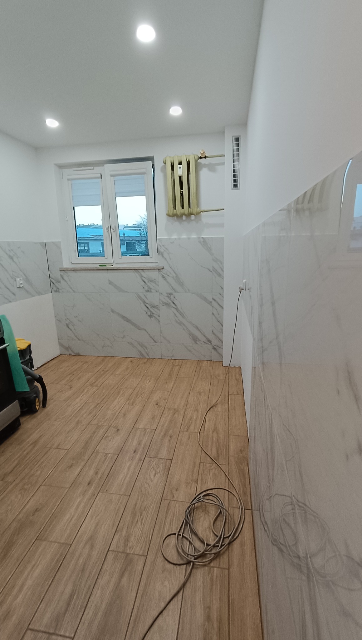 Wykończone wnętrze po remoncie: marmurowe płytki na ścianach do połowy wysokości, drewnopodobne panele na podłodze, okno i grzejnik. Widoczny odkurzacz.