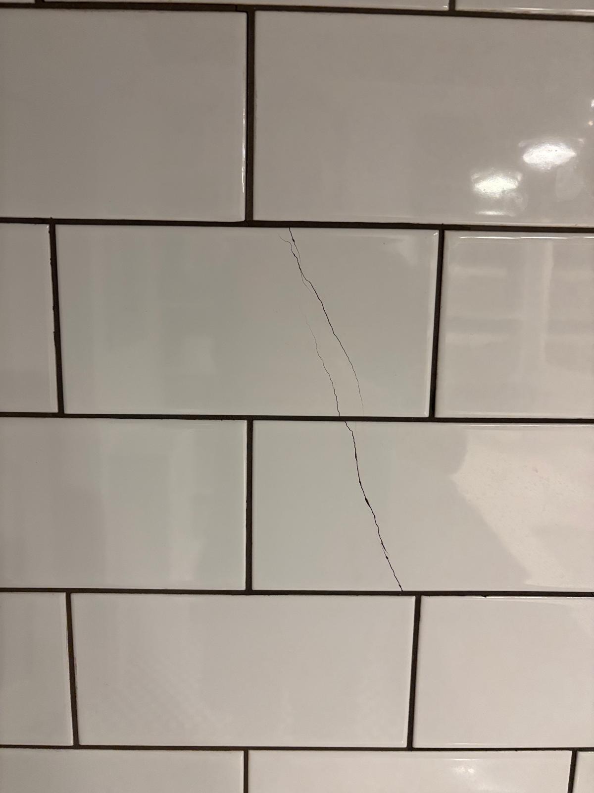 Białe, prostokątne płytki z ciemną fugą. Jedna z płytek ma pęknięcie biegnące przez jej środek, od góry do dołu. Widoczne odbicie światła na powierzchni płytek.