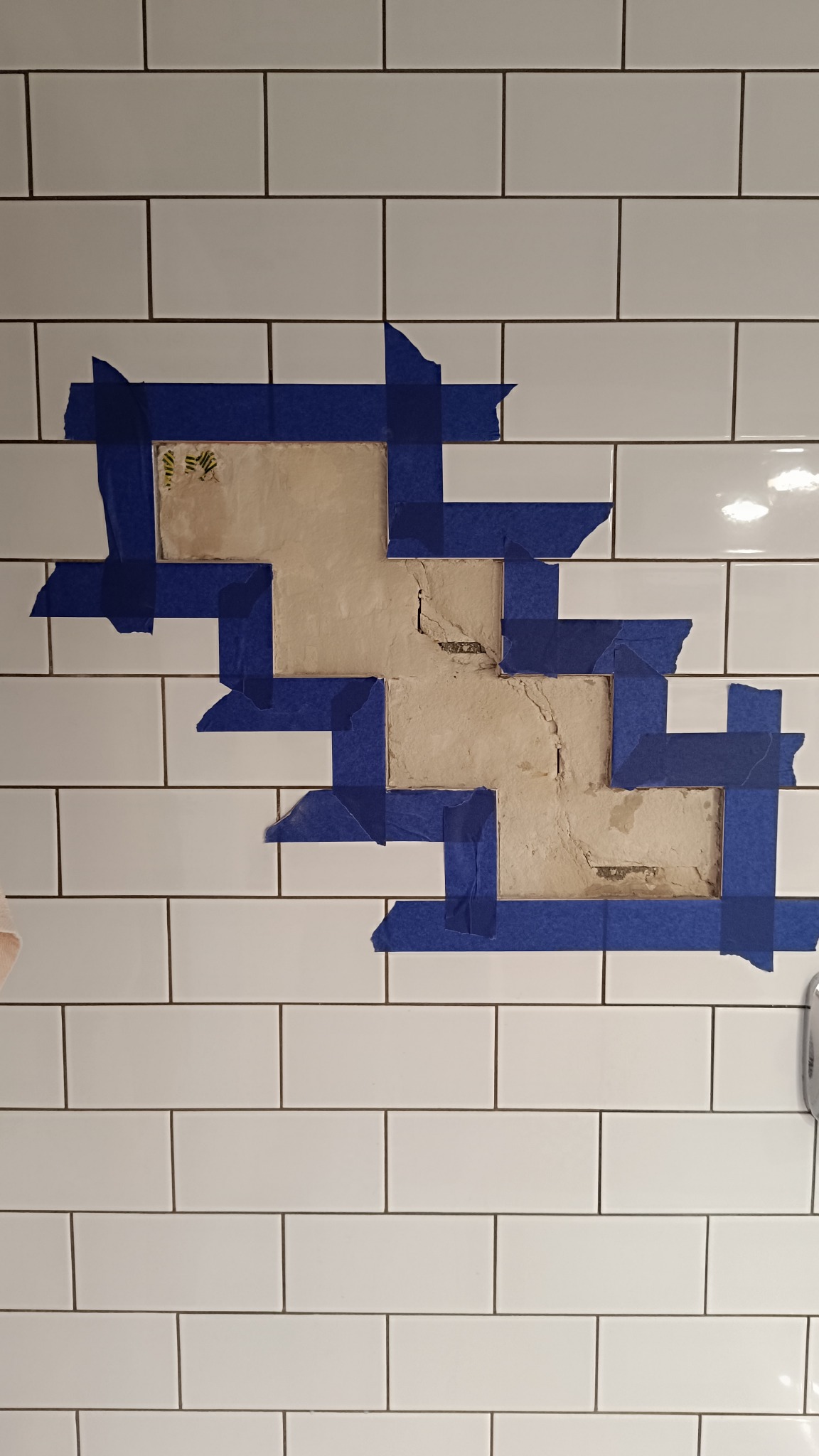 Białe płytki ścienne z uszkodzonym fragmentem, zabezpieczonym niebieską taśmą malarską przed naprawą. Widoczne pęknięcia i ubytki w podłożu.