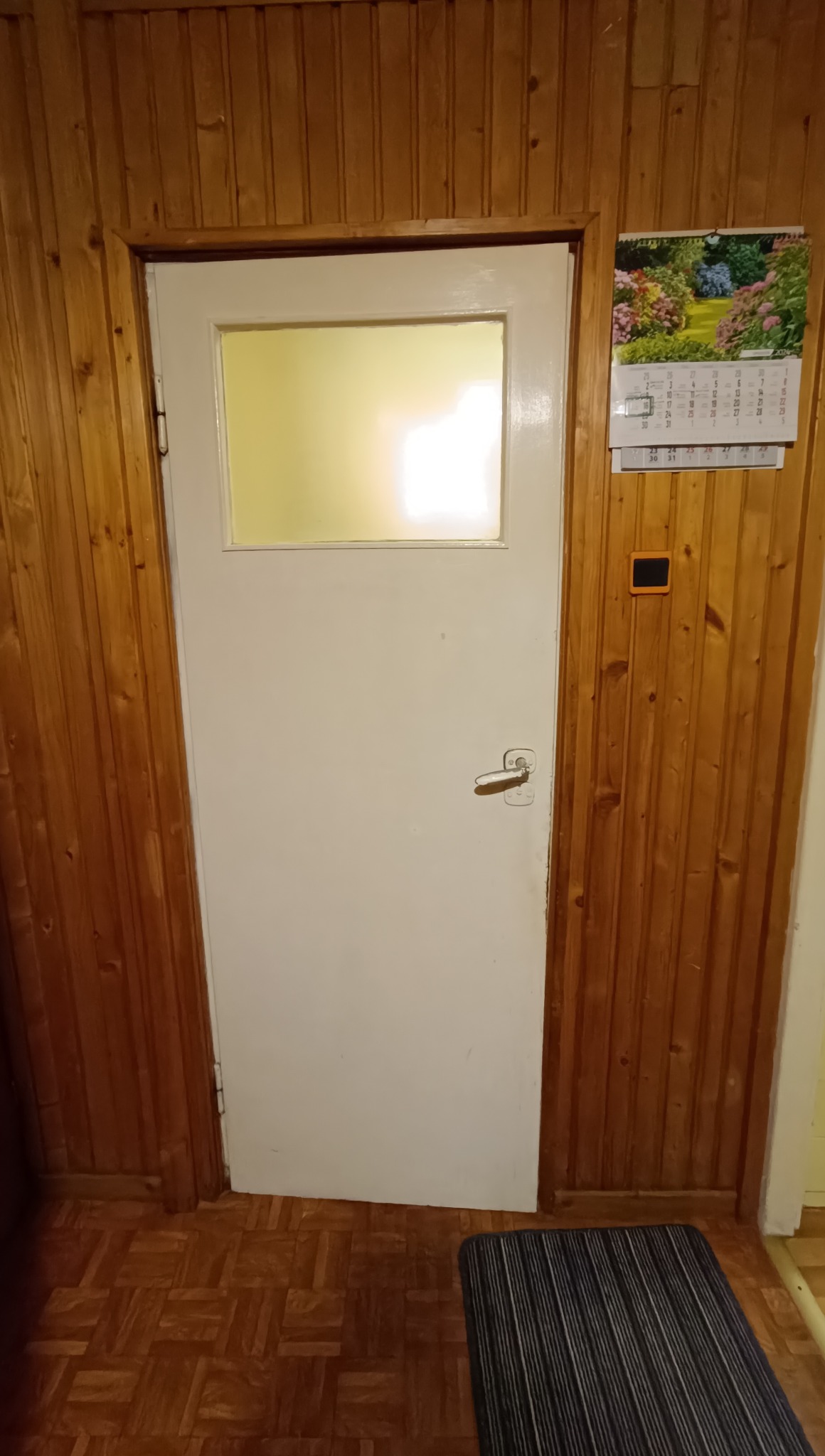 Białe drzwi z szybą w drewnianej boazerii, z kalendarzem na ścianie. Widoczny fragment dywanika i podłogi z wzorem parkietu. Oświetlenie wnętrza za drzwiami.
