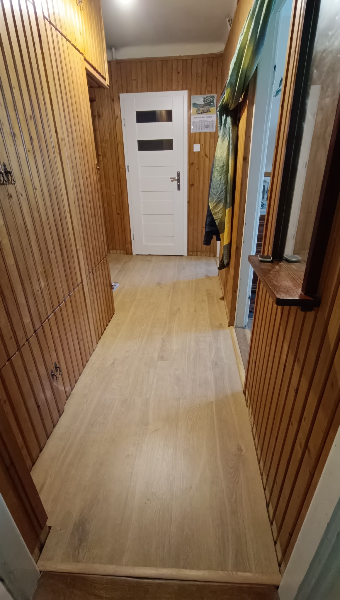 Jasny korytarz po remoncie z panelami podłogowymi i boazerią. Białe drzwi z przeszkleniami i drewniane wykończenia. Widok na wprost, ciepłe światło.