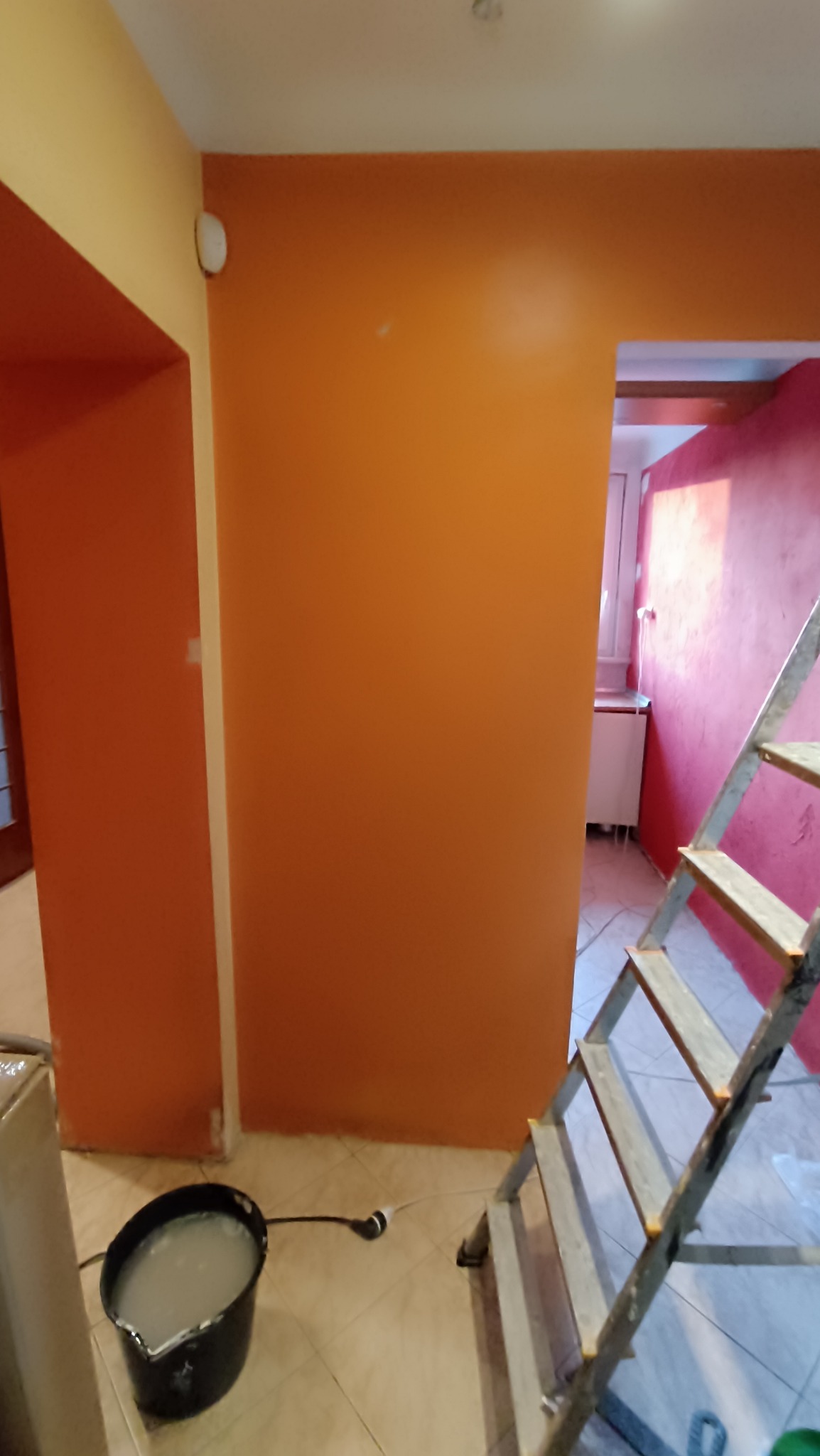 Pomarańczowa ściana po malowaniu, widoczna drabina i wiadro z farbą. W tle fragment różowej ściany w innym pomieszczeniu. Jasne płytki na podłodze.