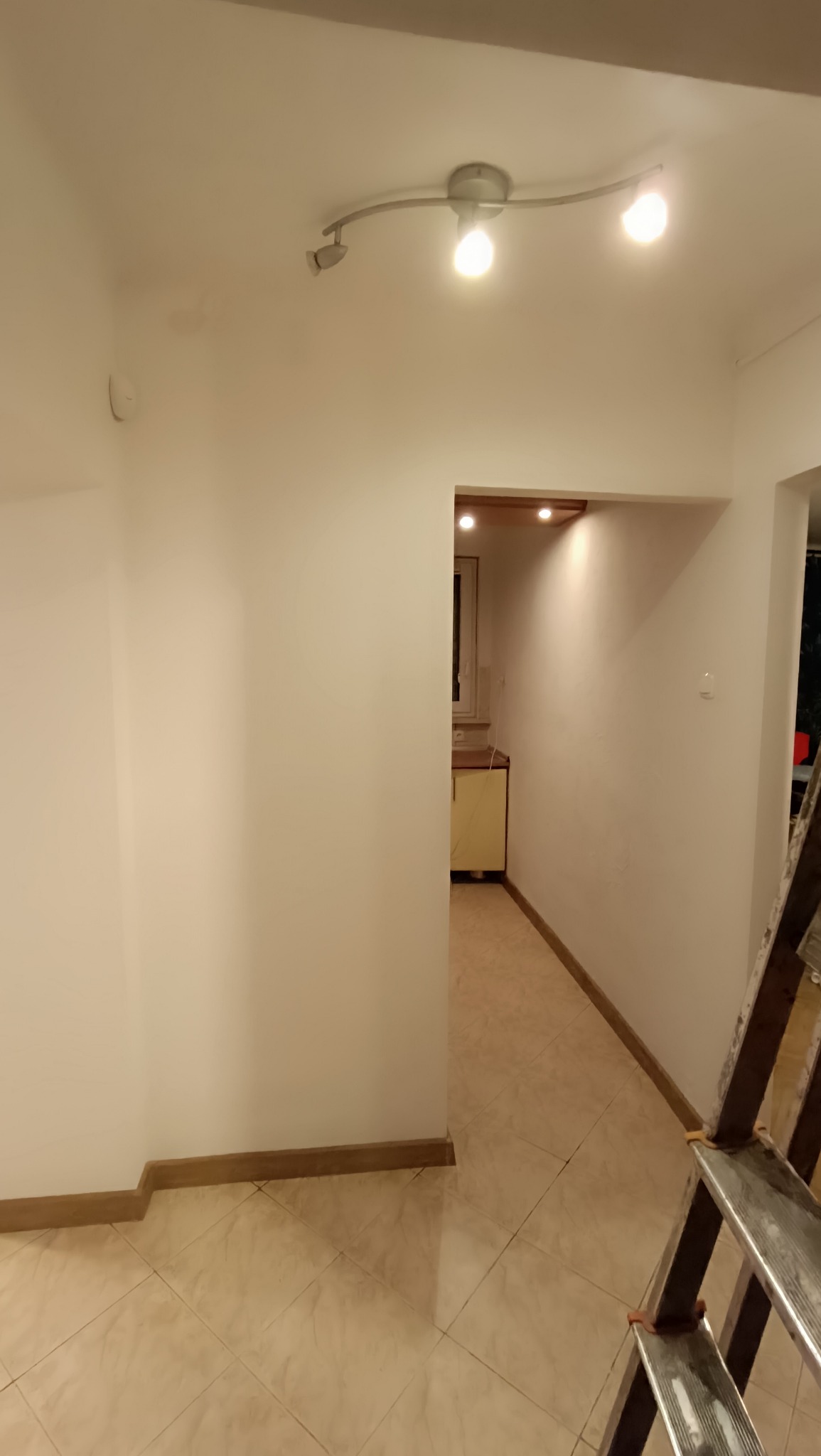 Wnętrze po malowaniu: białe ściany, beżowa podłoga z płytek, listwy przypodłogowe i lampa sufitowa. W tle widoczny fragment kuchni i drabina malarska.