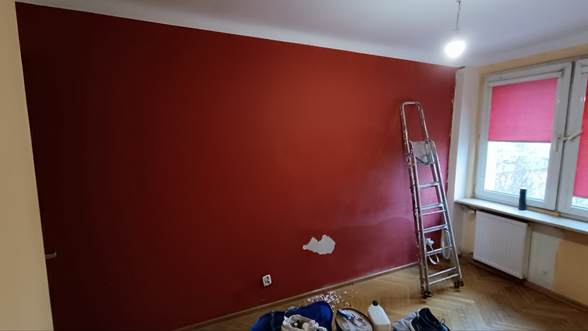 Czerwona ściana w trakcie malowania, widoczne niedomalowane fragmenty i drabina. Podłoga zabezpieczona, widoczne narzędzia malarskie. Okno z roletami w tle.