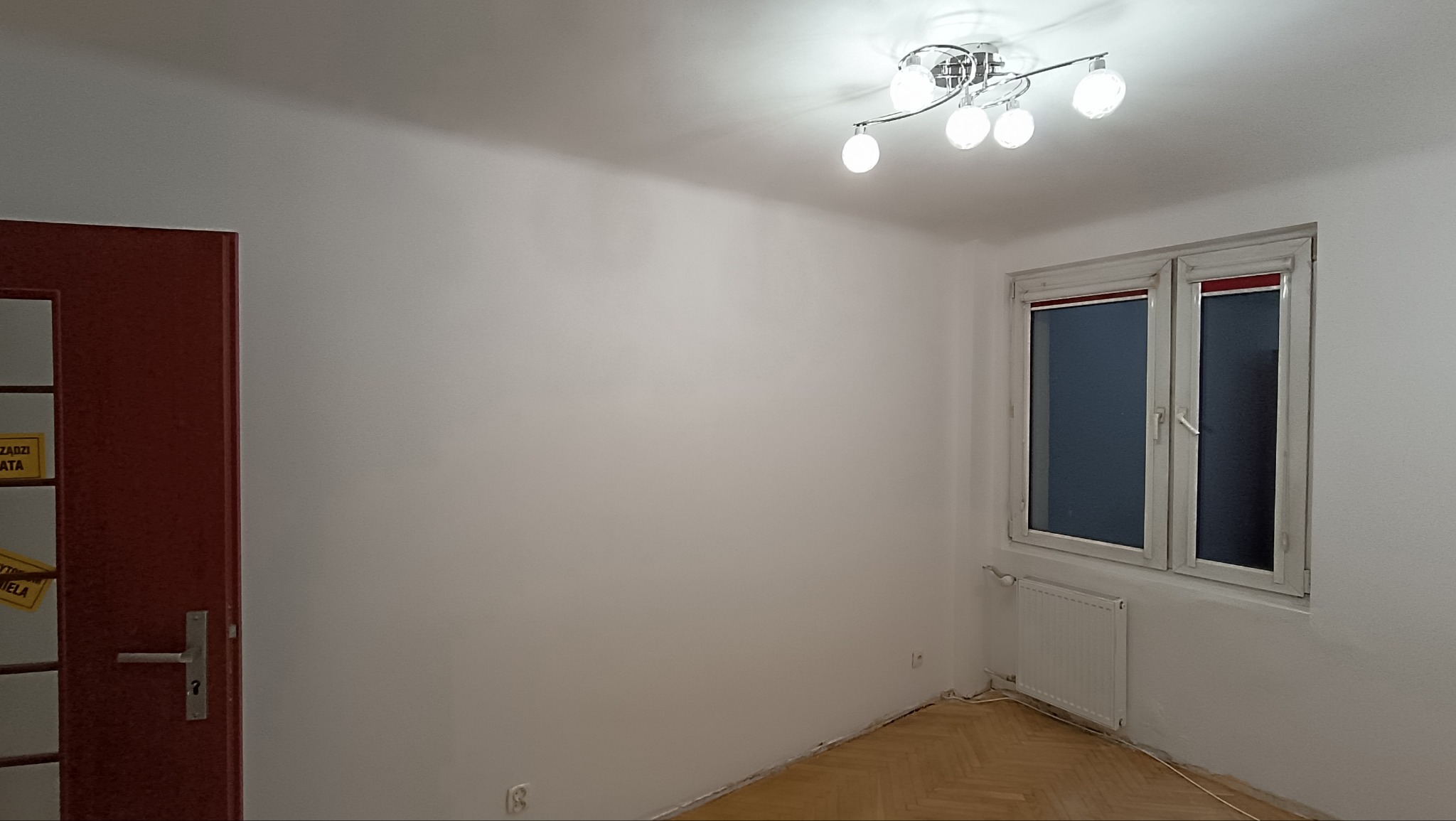 Jasne wnętrze po odświeżeniu: białe ściany, parkiet w jodełkę, okno z białymi ramami i czerwonym akcentem, nowoczesna lampa sufitowa. Widoczna część drzwi z żółtą taśmą.