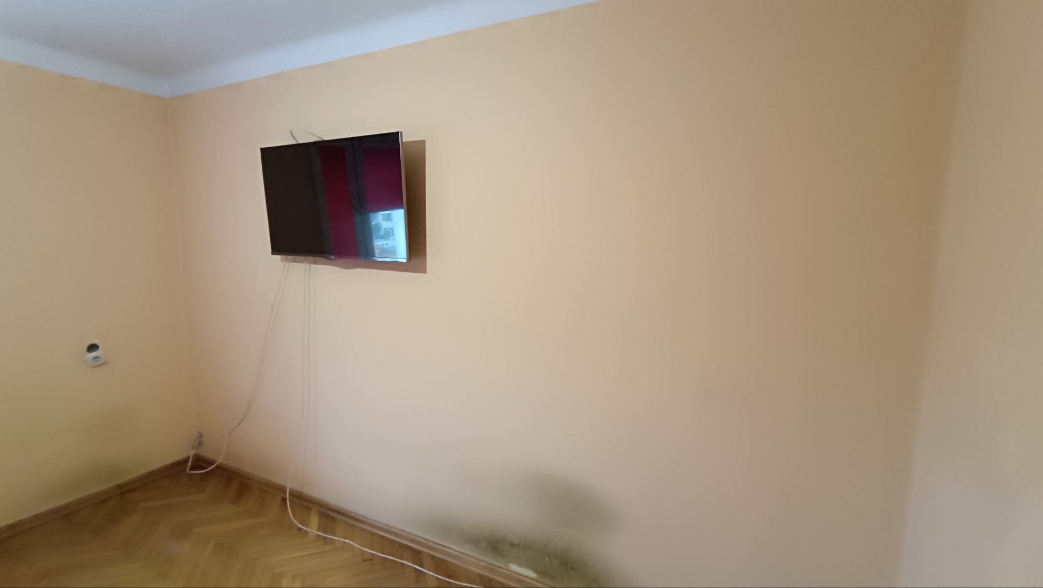 Pokój z telewizorem na ścianie w kolorze brzoskwiniowym. Widoczne kable, gniazdko elektryczne i parkiet. Na dole ściany widoczne ślady wilgoci.