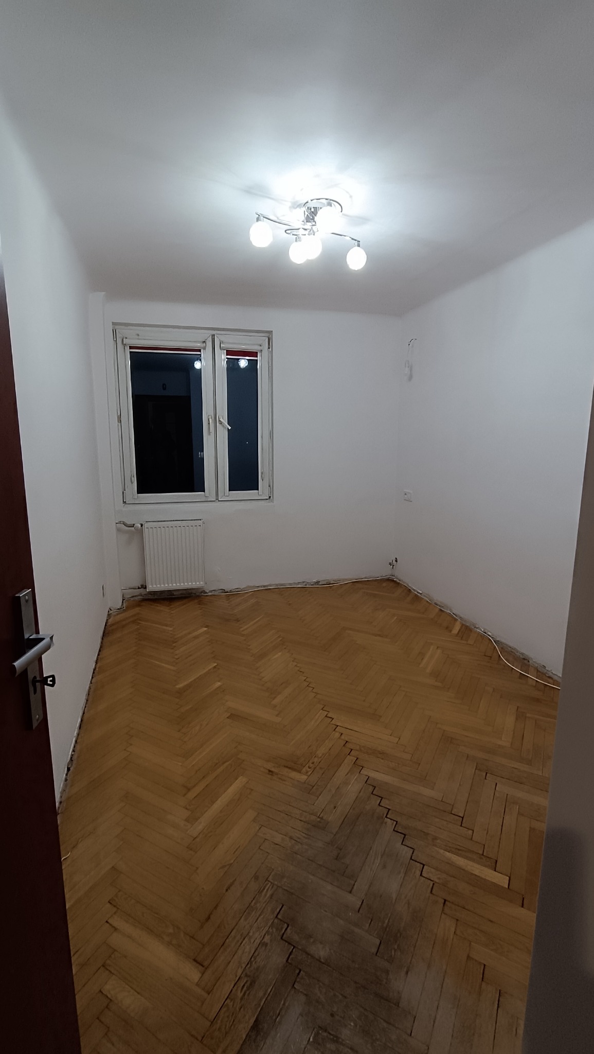 Pusty pokój z parkietem ułożonym w jodełkę, białymi ścianami i oknem. Widoczny grzejnik, lampa sufitowa i fragment otwartych drzwi. Przygotowanie do remontu.