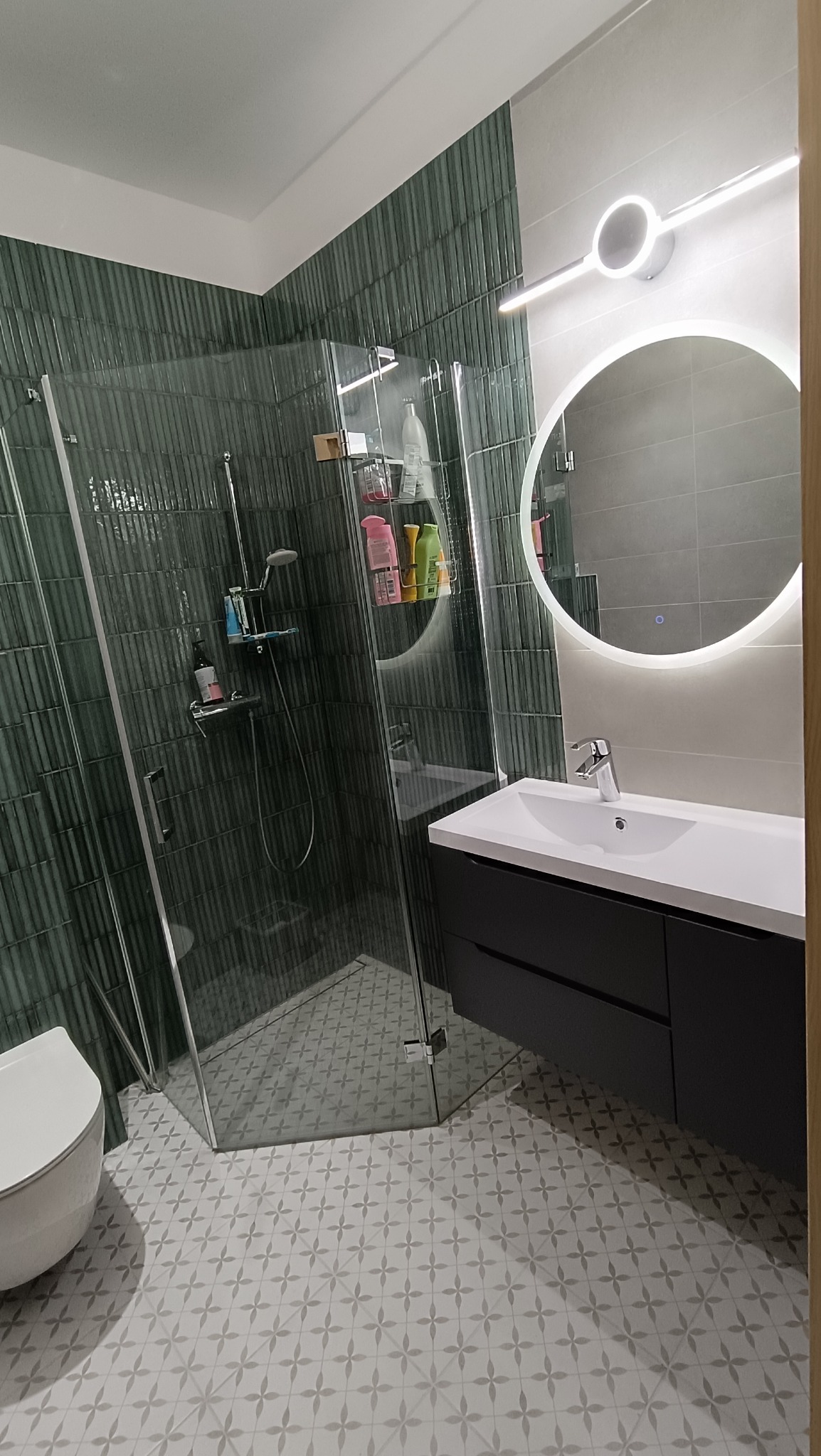 Nowoczesna łazienka z kabiną prysznicową typu walk-in, umywalką z szafką w kolorze grafitowym i okrągłym lustrem z podświetleniem LED. Na ścianach zielone płytki.