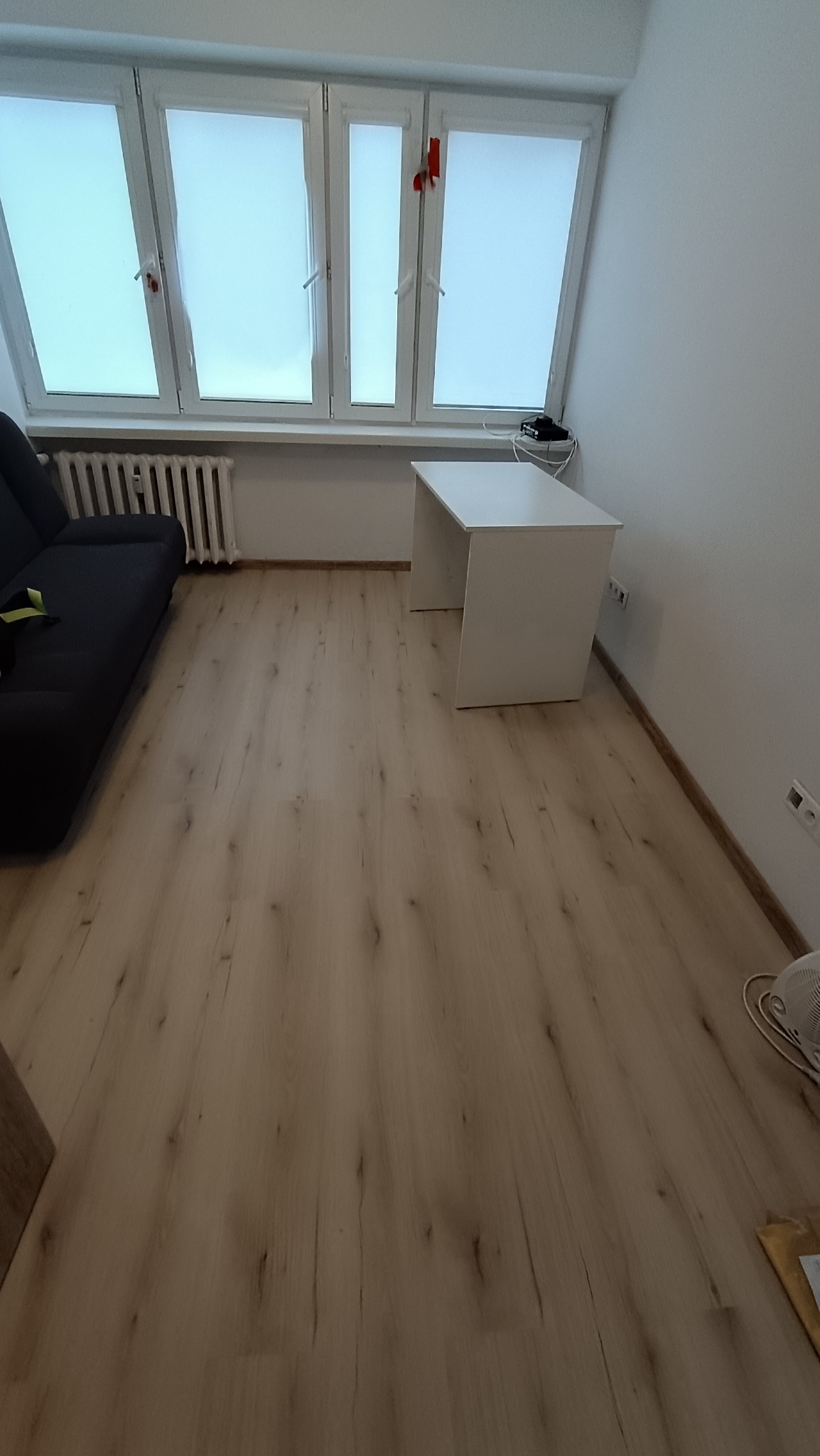 Jasne panele podłogowe w pokoju z szarą sofą i białym biurkiem pod oknem. Widoczne listwy przypodłogowe i grzejnik. Minimalistyczne wnętrze po remoncie.