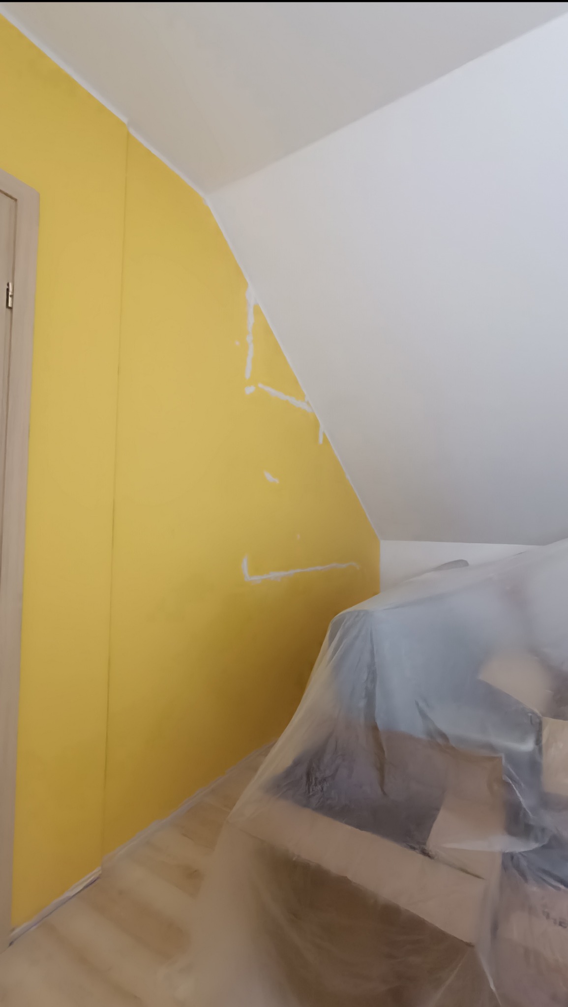 Żółta ściana w pokoju na poddaszu, częściowo zagruntowana, z widocznym zabezpieczeniem mebli folią malarską. Przygotowania do malowania.