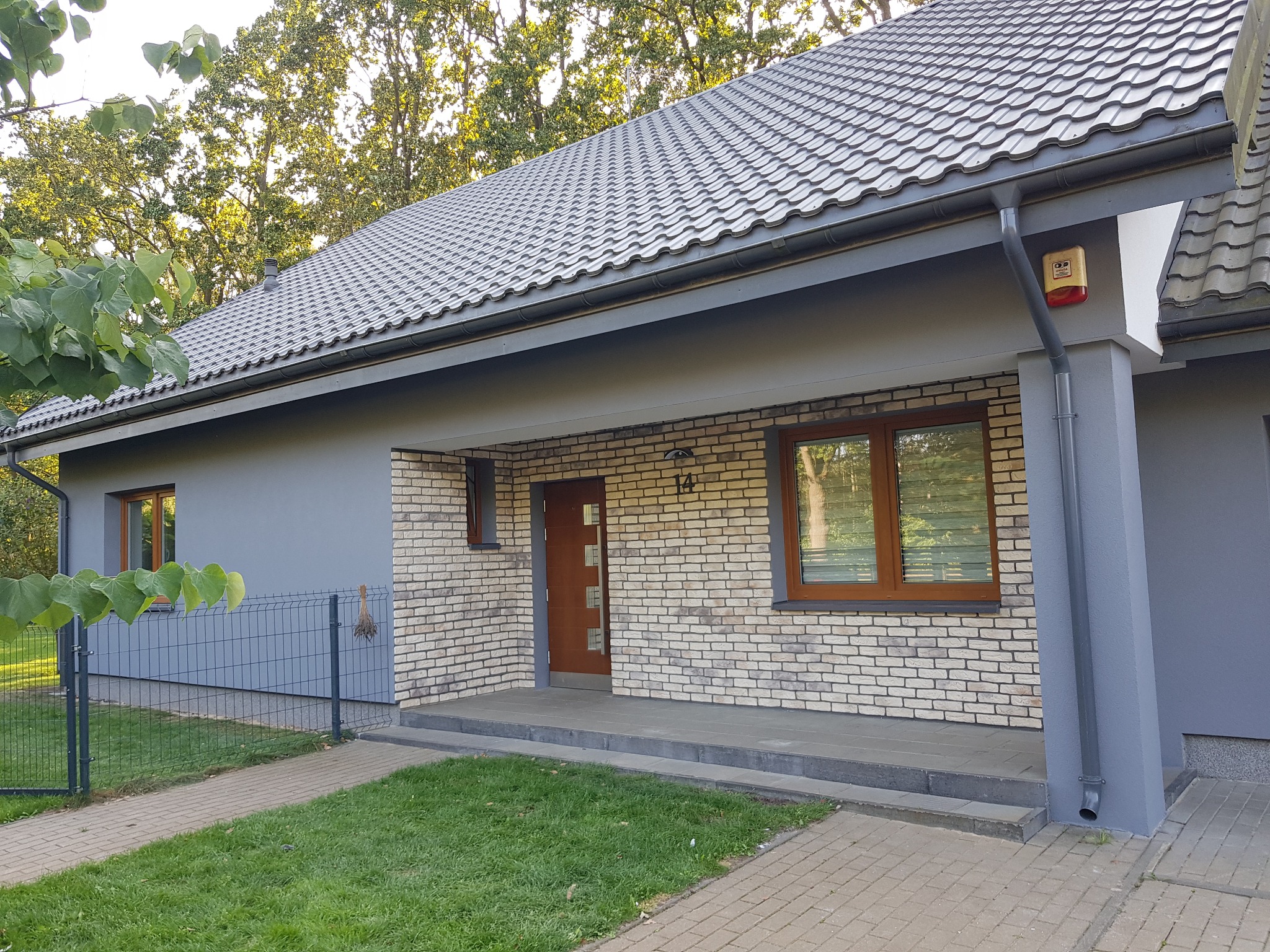 Szary dom jednorodzinny z ceglaną elewacją, drewnianymi oknami i metalową dachówką. Widoczny fragment ogrodu i chodnika. Alarm na ścianie.