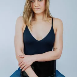 Portret kobiety siedzącej na krześle, ubrana w czarny top na ramiączkach i dżinsy, z naturalnym makijażem i tatuażem na ramieniu, na jasnym tle.