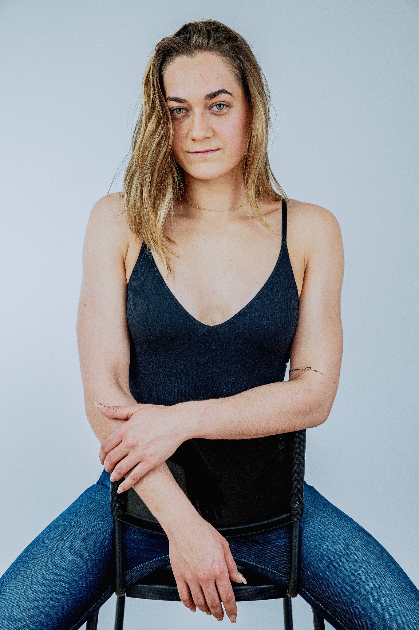 Portret kobiety siedzącej na krześle, ubrana w czarny top na ramiączkach i dżinsy, z naturalnym makijażem i tatuażem na ramieniu, na jasnym tle.