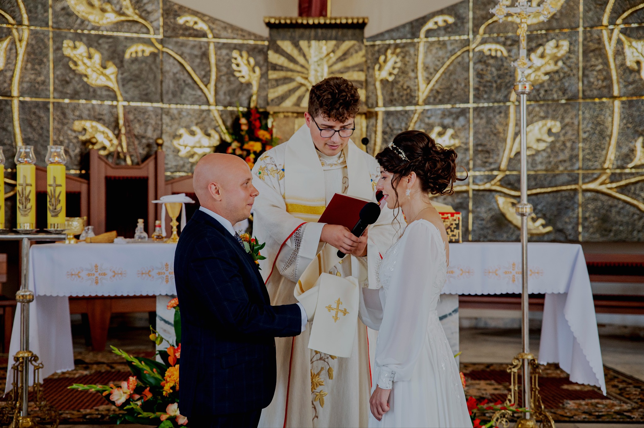 Uroczystość ślubna w kościele: para młoda podczas przysięgi małżeńskiej, ksiądz z mikrofonem, dekoracje kwiatowe i złote ornamenty na ścianie za ołtarzem.