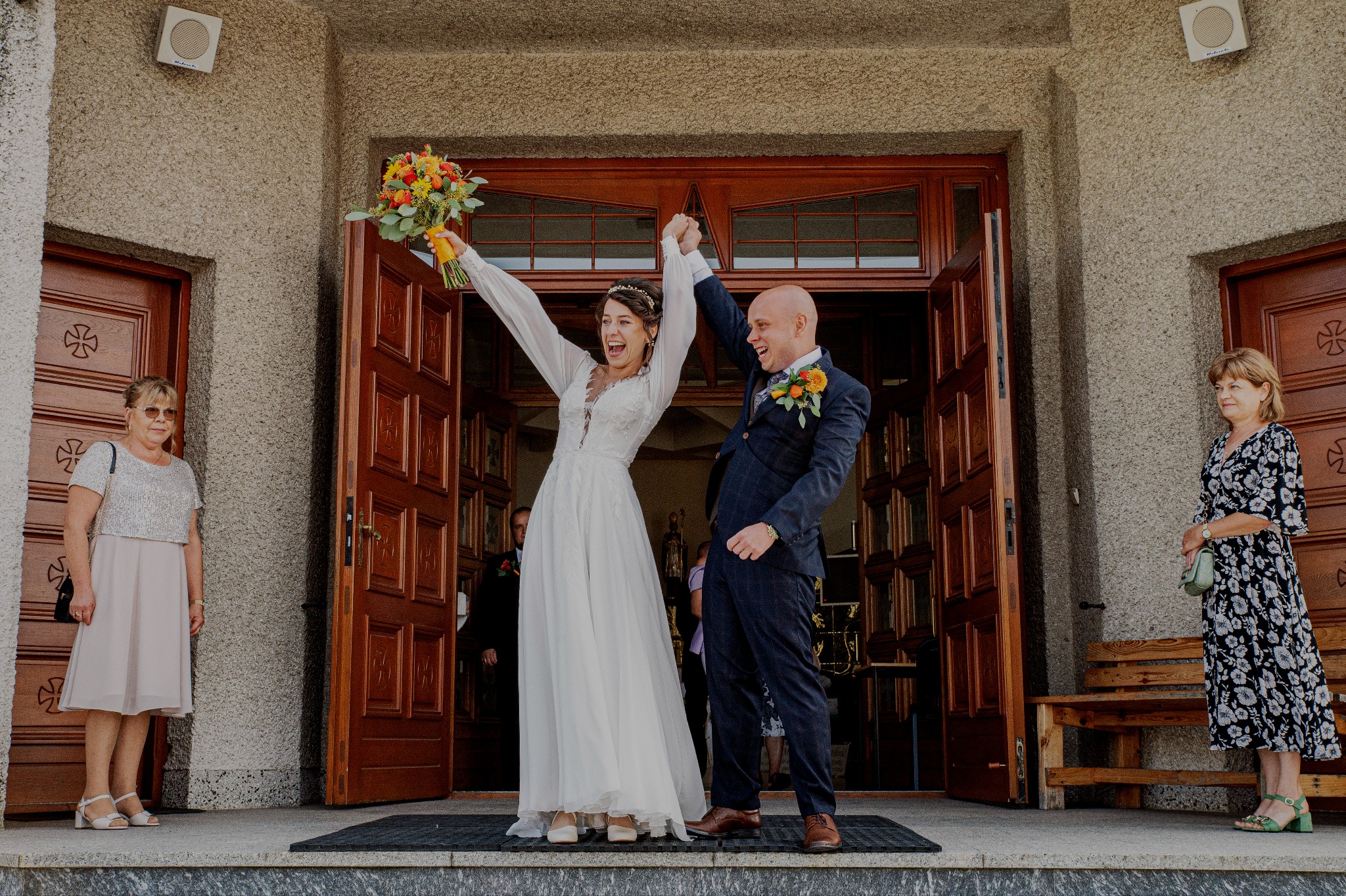 Uśmiechnięta para młoda, trzymając się za ręce, wychodzi z kościoła. Panna młoda unosi bukiet kwiatów w górę. Po bokach stoją kobiety.