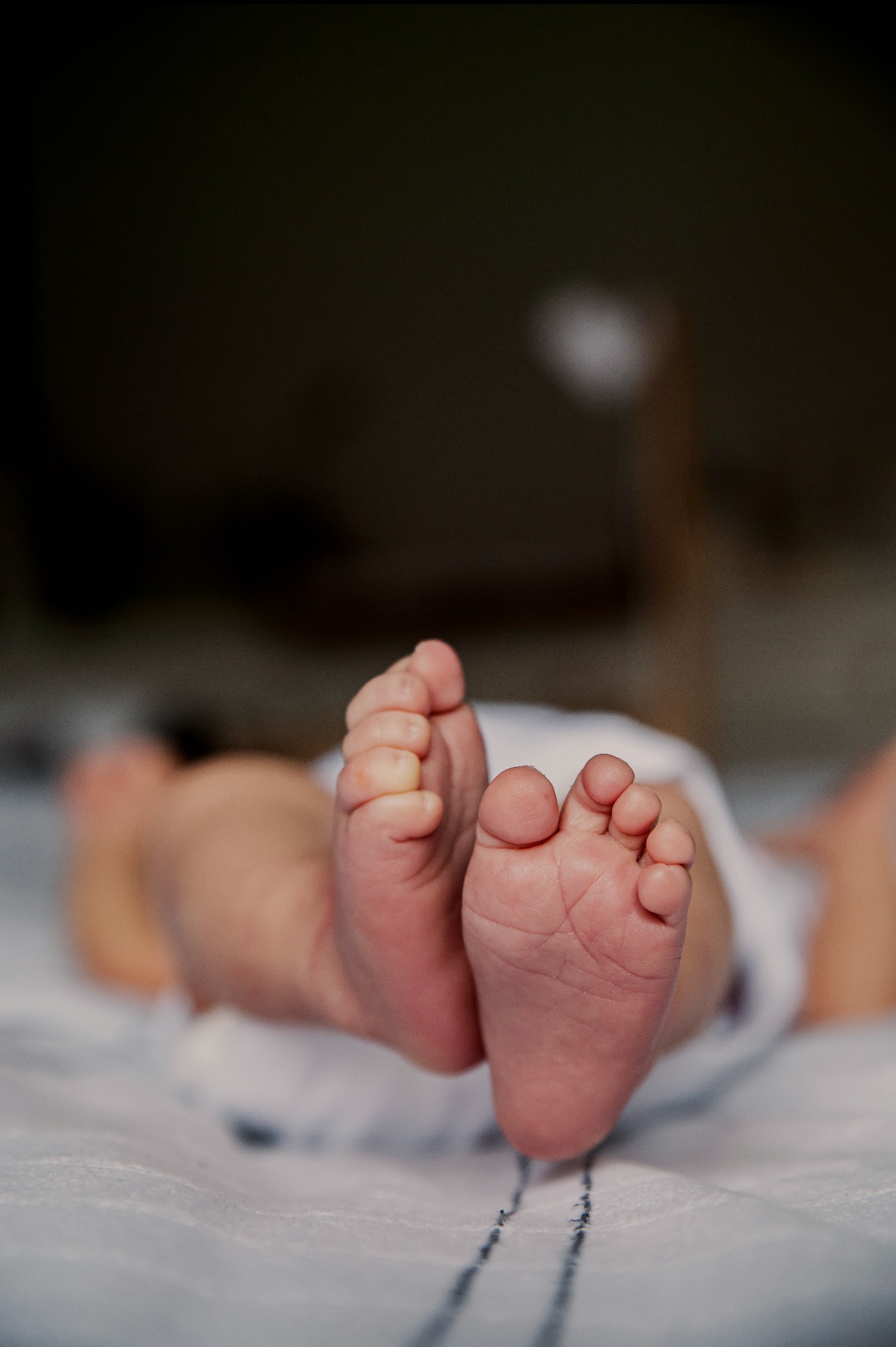 Drobne stópki noworodka na białym, wzorzystym kocyku, uchwycone w zbliżeniu z góry. Delikatne światło podkreśla fałdki skóry i małe paluszki.