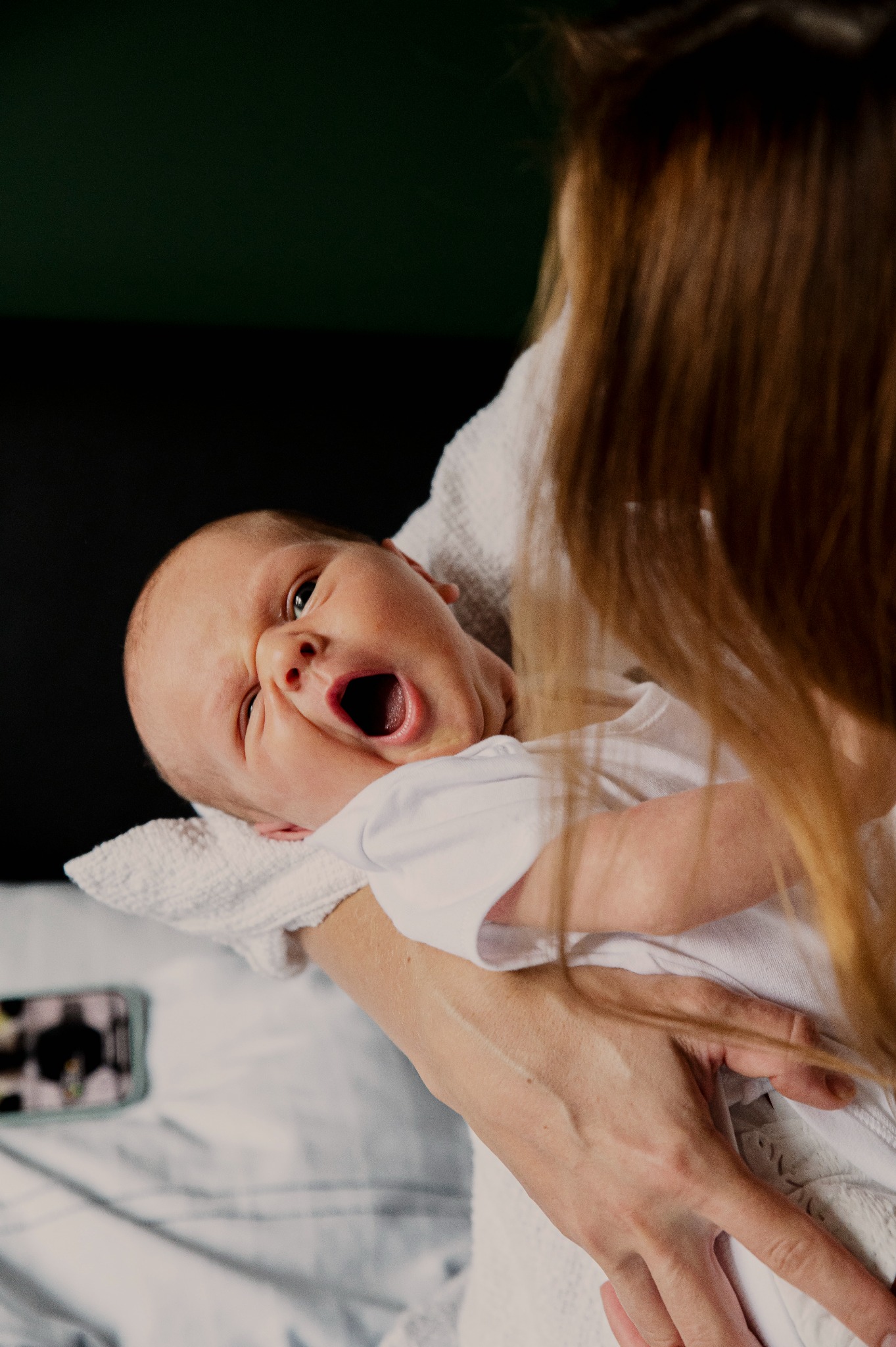 Noworodek z otwartymi ustami, trzymany na rękach przez kobietę z rudymi włosami. W tle ciemna ściana i telefon na łóżku.