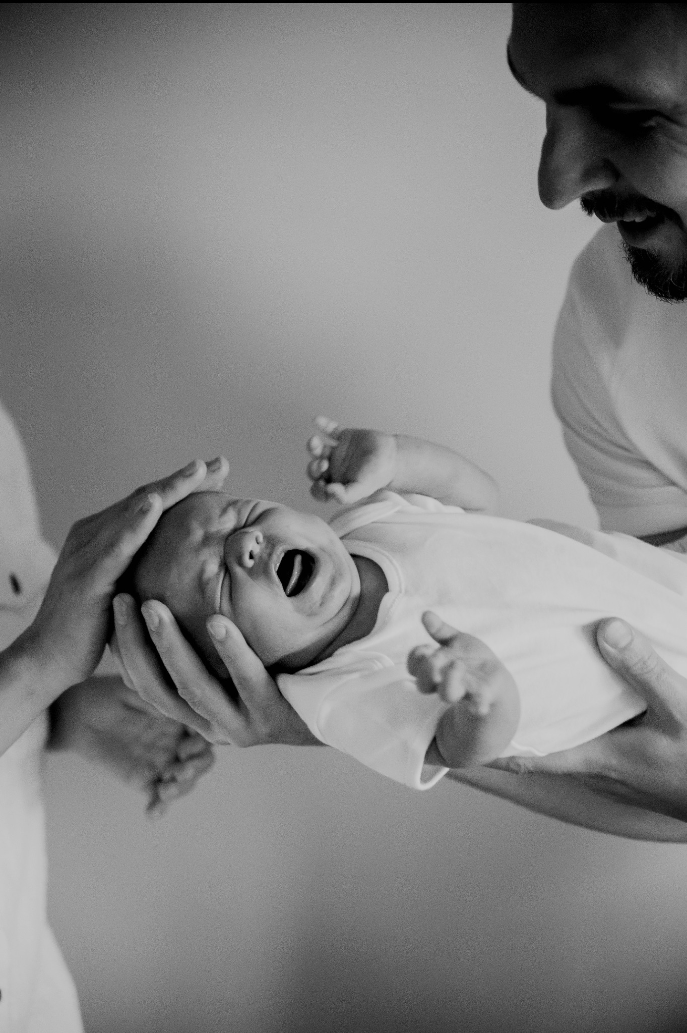 Czarno-białe zbliżenie: rodzice trzymają noworodka, który szeroko ziewa, ukazując język. Widoczna twarz ojca z brodą. Proste tło, miękkie światło.