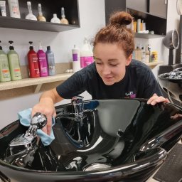 VIP CLINING Inha Bilous - Pracownica salonu fryzjerskiego czyści umywalkę do włosów, używając niebieskiej ściereczki. W tle półki z kosmetykami i czarne szafki.