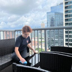 VIP CLINING Inha Bilous - Osoba czyści szklany stolik na balkonie z widokiem na panoramę Katowic. W tle wysokie budynki i pochmurne niebo.