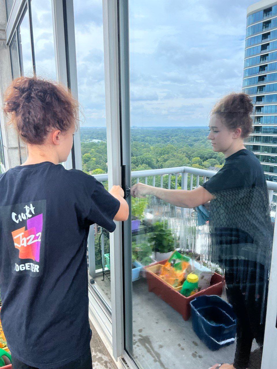 Osoba czyści szklane drzwi balkonowe z widokiem na zieleń miejską i wieżowiec, używając ściereczki i płynu, z bliska widoczny balkon z donicami.