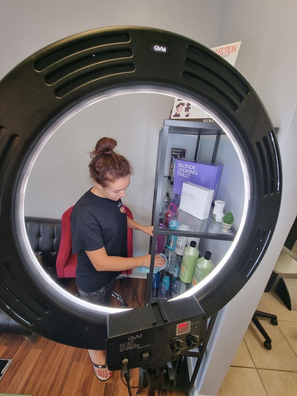 Kobieta przeciera półki w salonie kosmetycznym, oświetlona lampą pierścieniową GVM. Widoczne kosmetyki, regał i elementy wyposażenia wnętrza.