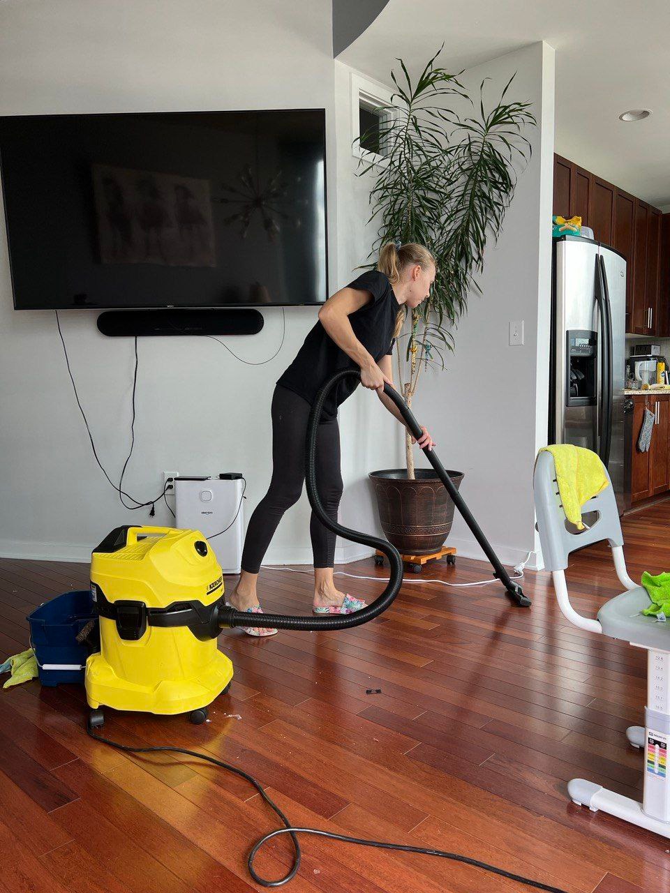 Kobieta odkurza parkiet odkurzaczem Karcher. W tle: telewizor, roślina doniczkowa, krzesełko dziecięce. Widoczne drewniane podłogi i jasne ściany.