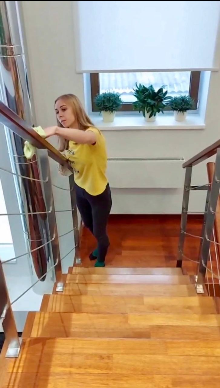 Kobieta w żółtej koszulce czyści drewniane schody z metalową balustradą. W tle okno z roślinami i biała roleta. Jasne wnętrze, widok z góry na schody.