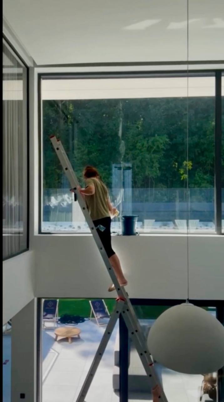 Kobieta na drabinie myje wysokie okno w nowoczesnym domu. Widoczny kubeł z wodą i ściereczka. Zza okna widok na zieleń. U dołu taras z meblami.