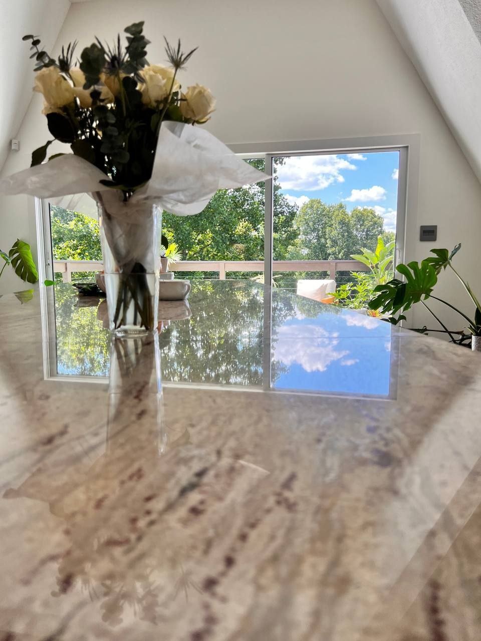 Błyszczący blat stołu odbija bukiet białych róż w wazonie i widok za oknem na zielone drzewa i błękitne niebo z chmurami w jasnym wnętrzu.