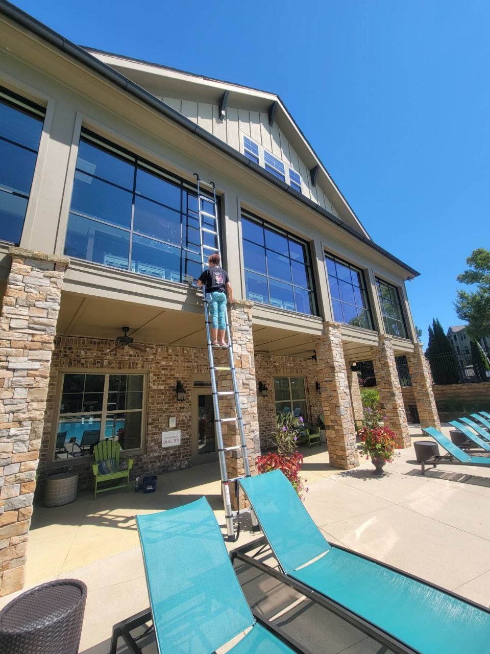 Osoba na drabinie myje duże okna budynku z kamienną elewacją, widok z dołu, błękitne niebo, leżaki w tle. Usługa mycia okien w obiekcie.