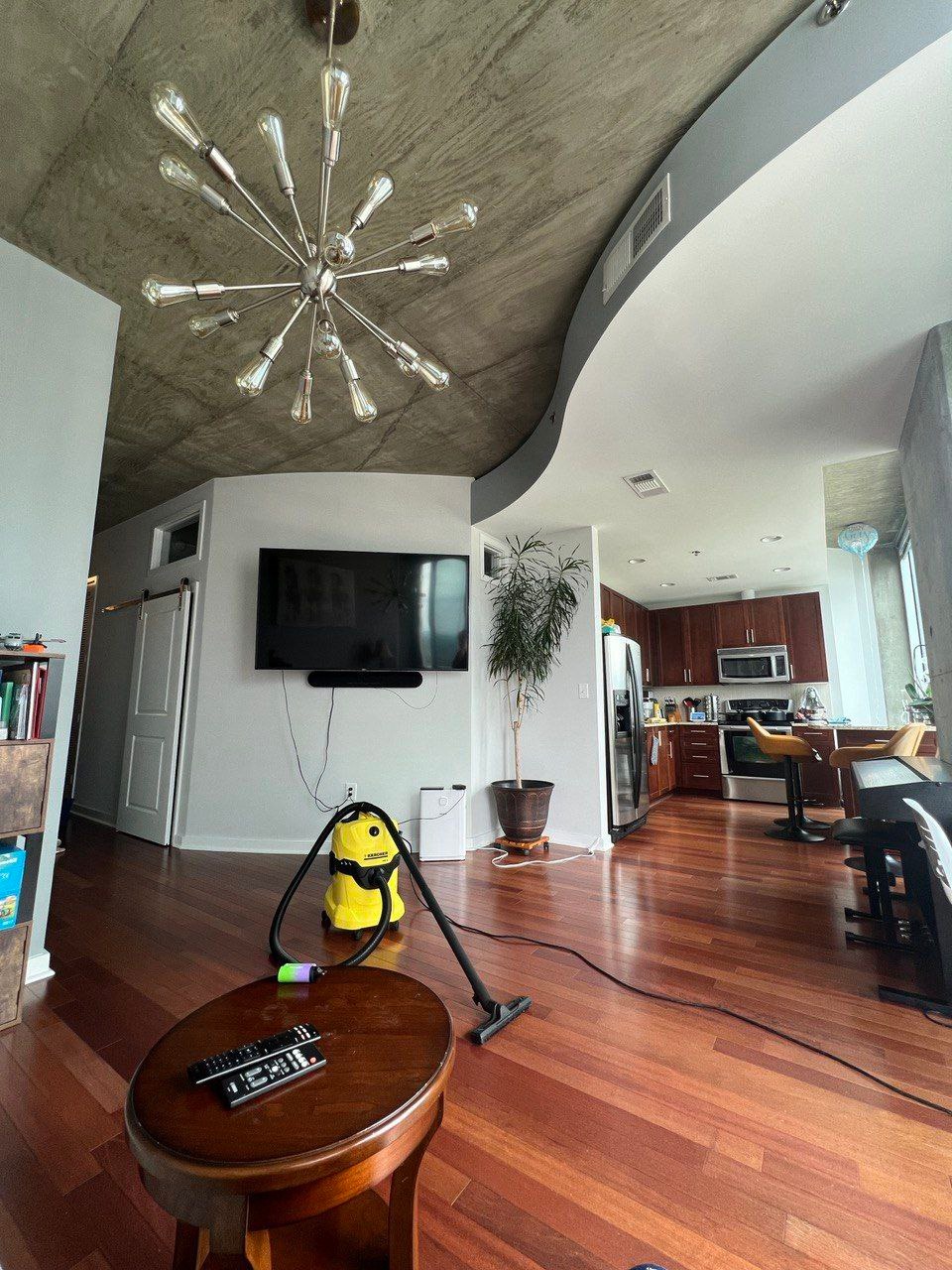 Żółty odkurzacz Karcher na drewnianej podłodze w nowoczesnym salonie z betonowym sufitem i designerską lampą. Widok na kuchnię w tle.