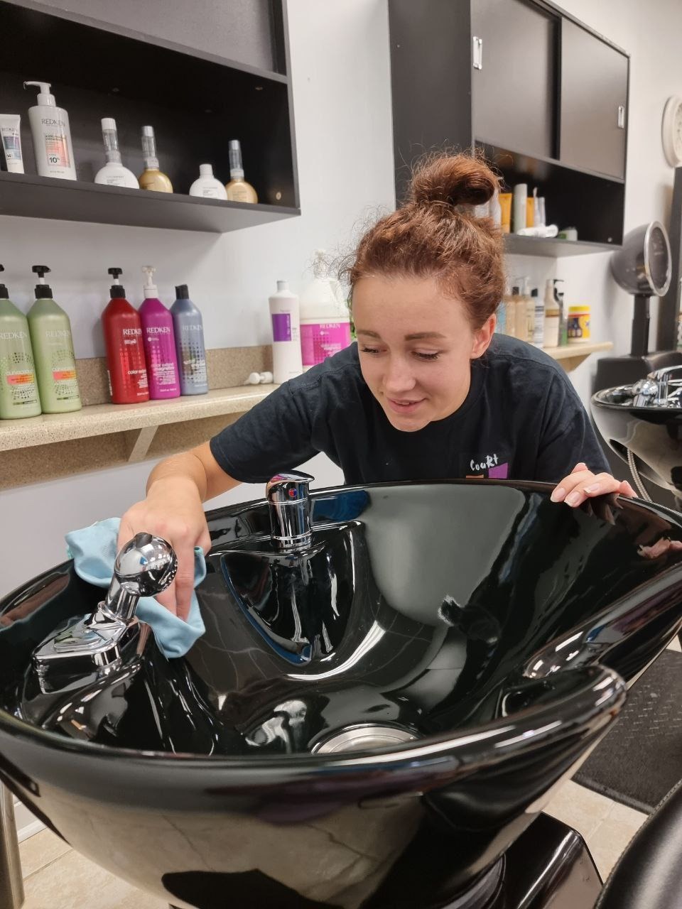 Pracownica salonu fryzjerskiego czyści umywalkę do włosów, używając niebieskiej ściereczki. W tle półki z kosmetykami i czarne szafki.