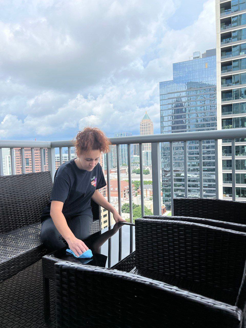 Osoba czyści szklany stolik na balkonie z widokiem na panoramę Katowic. W tle wysokie budynki i pochmurne niebo.