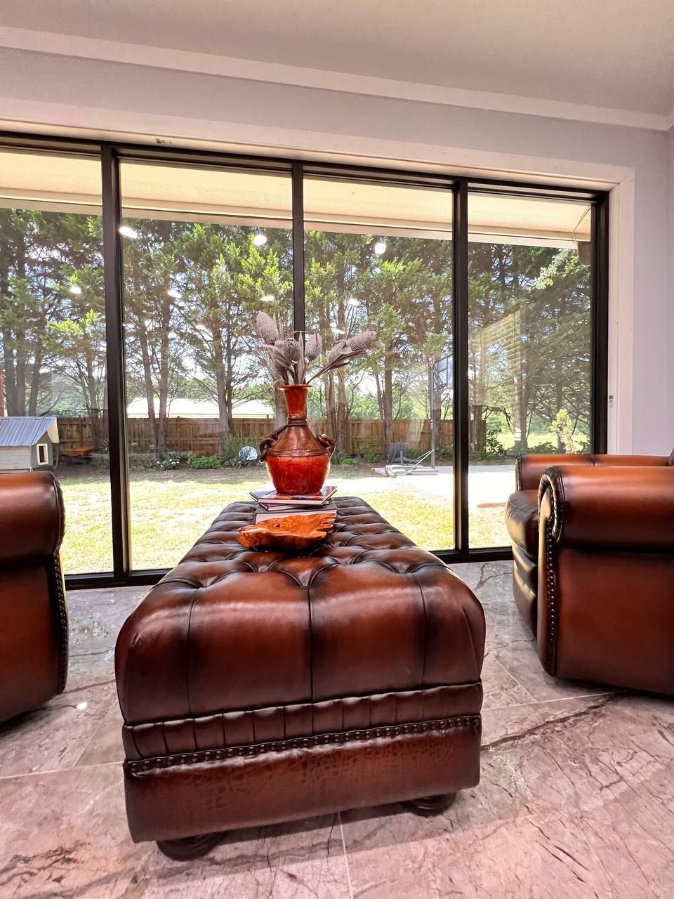 Elegancki salon z skórzanym pufem, fotelami i widokiem na ogród przez panoramiczne okno. Wazon z suszem na pufie, marmurowa podłoga.