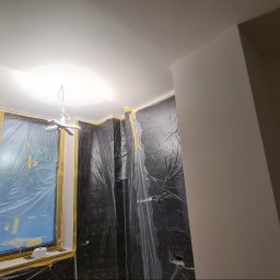 Remonty Wojciech - Pokój zabezpieczony folią malarską i taśmą przed malowaniem. Widoczne okno z zabezpieczeniem, sufit i fragment pomalowanej ściany. Przygotowanie do malowania.