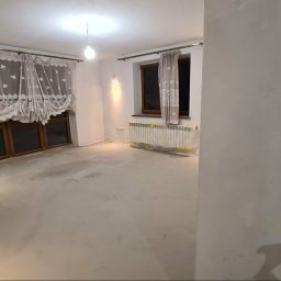 Remonty Wojciech - Pokój po wylewce, przygotowany do dalszych prac wykończeniowych. Okna z drewnianymi ramami, zasłony z koronkowym wzorem. Grzejnik zabezpieczony folią.