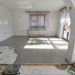 Remonty Wojciech - Wnętrze w trakcie wylewania samopoziomującej wylewki, widoczne worki z zaprawą, narzędzia i fragmenty nierównej powierzchni. Jasne światło dzienne.