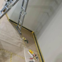 Remonty Wojciech - Świeżo pomalowane jasnoszare ściany klatki schodowej zabezpieczone taśmą malarską, z rozstawioną drabiną i narzędziami malarskimi na podłodze.