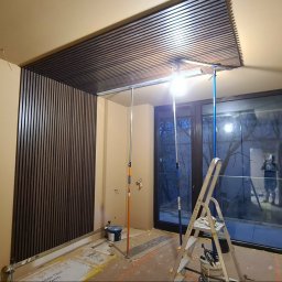 Remonty Wojciech - Montaż paneli dekoracyjnych na ścianie i suficie w trakcie wykańczania wnętrza. Widoczne narzędzia, drabina i oświetlenie budowlane. Ściany w kolorze beżowym.