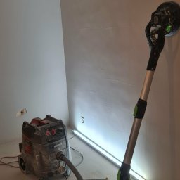 Remonty Wojciech - Ściana po szpachlowaniu, szlifierka do gipsu z odkurzaczem przemysłowym. Widoczne ślady po gładzi, lampa oświetleniowa. Przygotowanie do malowania.