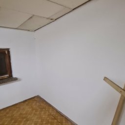Remonty Wojciech - Wnętrze pokoju po malowaniu ścian na biało, widoczna podłoga w brązową kratę i fragment drewnianej drabiny oparty o ścianę. Sufit wykończony panelami.