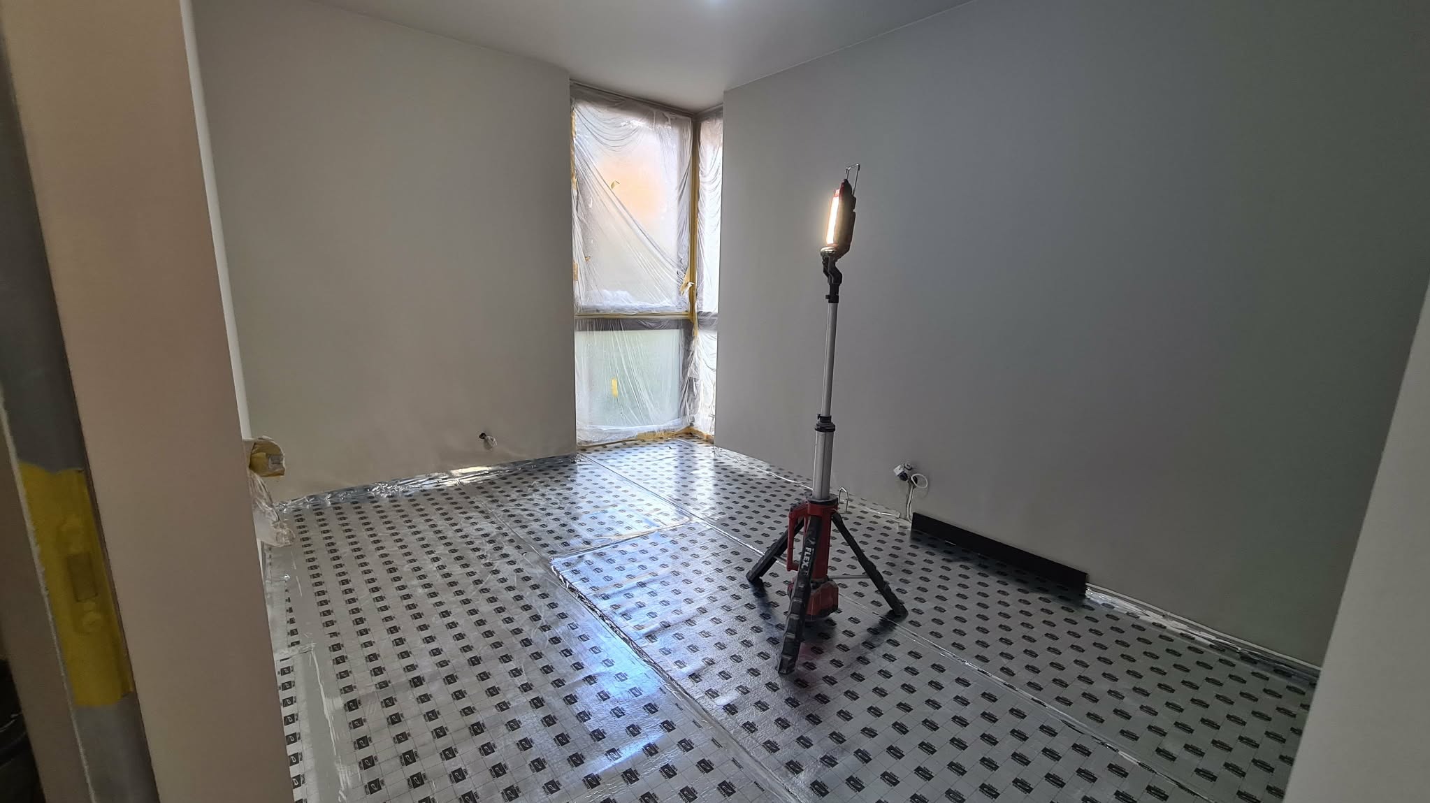 Pokój w trakcie remontu: przygotowane podłoże pod ogrzewanie podłogowe, ściany po szpachlowaniu, okno zabezpieczone folią, lampa budowlana oświetla wnętrze.