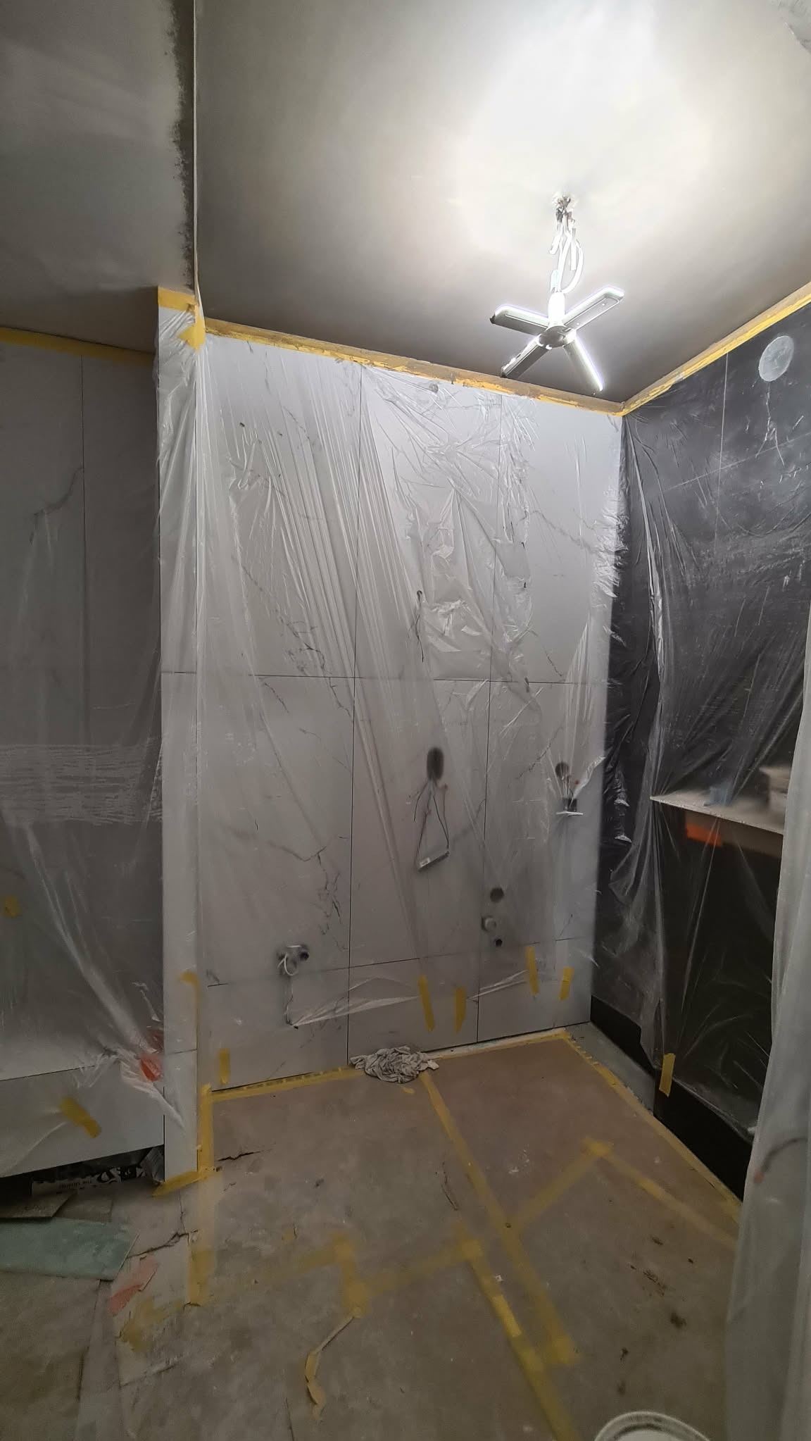 Łazienka w trakcie remontu: ściany wyłożone płytkami imitującymi marmur, zabezpieczone folią malarską. Widoczne przygotowane instalacje wodne i fragment sufitu z lampą.