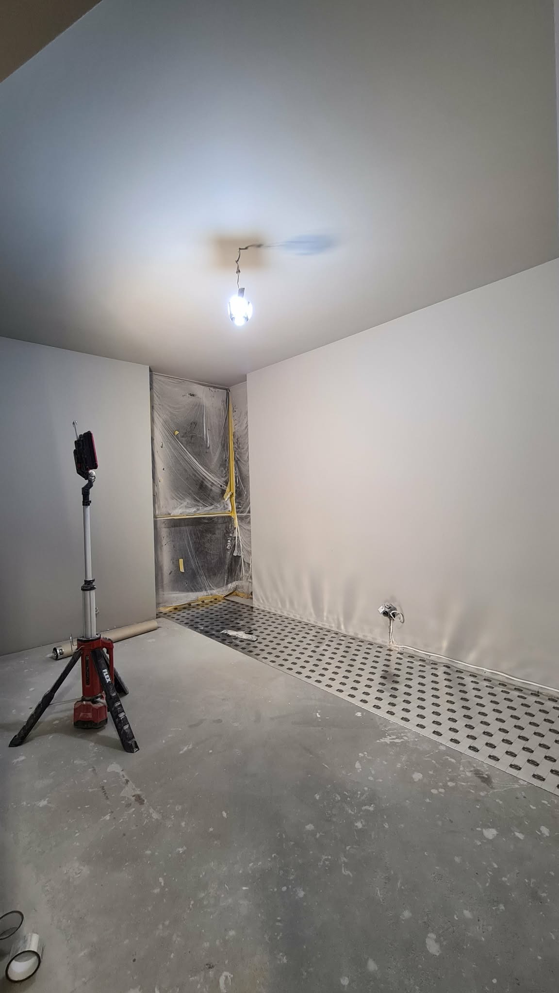 Wnętrze w trakcie remontu: gładkie, białe ściany, zabezpieczone folią drzwi, podłoga z wylewką i kratką dylatacyjną. Na pierwszym planie lampa budowlana.