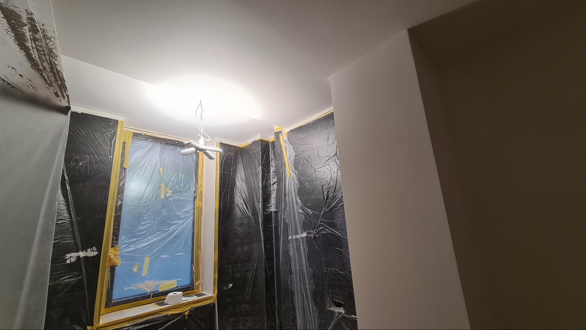 Pokój zabezpieczony folią malarską i taśmą przed malowaniem. Widoczne okno z zabezpieczeniem, sufit i fragment pomalowanej ściany. Przygotowanie do malowania.