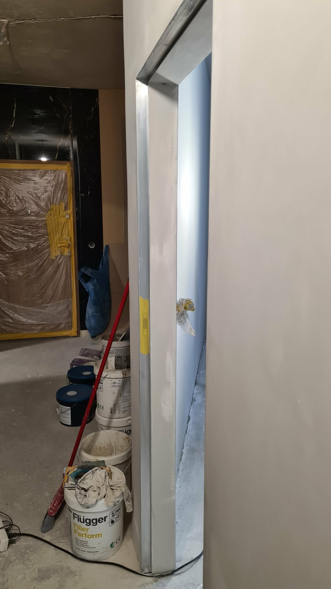 Przygotowanie do malowania: ościeżnica po szpachlowaniu, widoczne puszki z farbą Flugger, zabezpieczone drzwi w tle. Etap prac wykończeniowych.